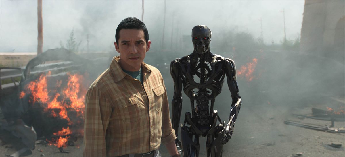 Gabriel Luna and a Terminator exoskeleton stand on a bridge in front of a burning car fuming diesel smoke