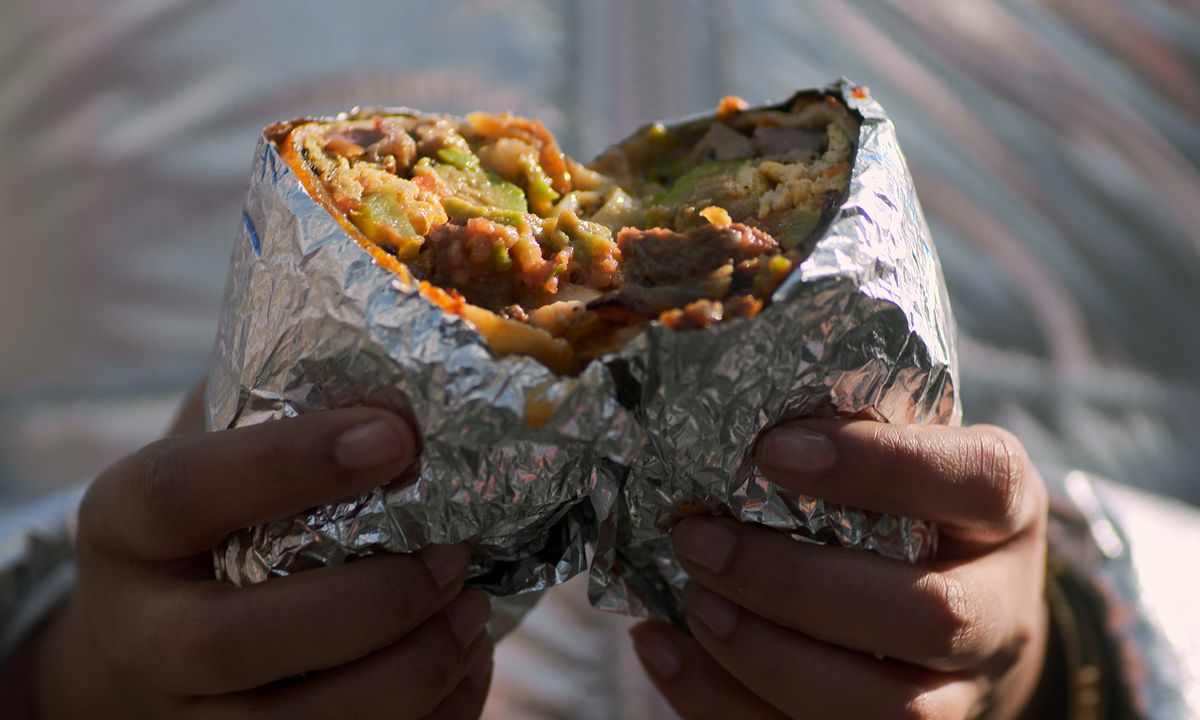 Two hands hold a foil wrapped burrito being torn in half.