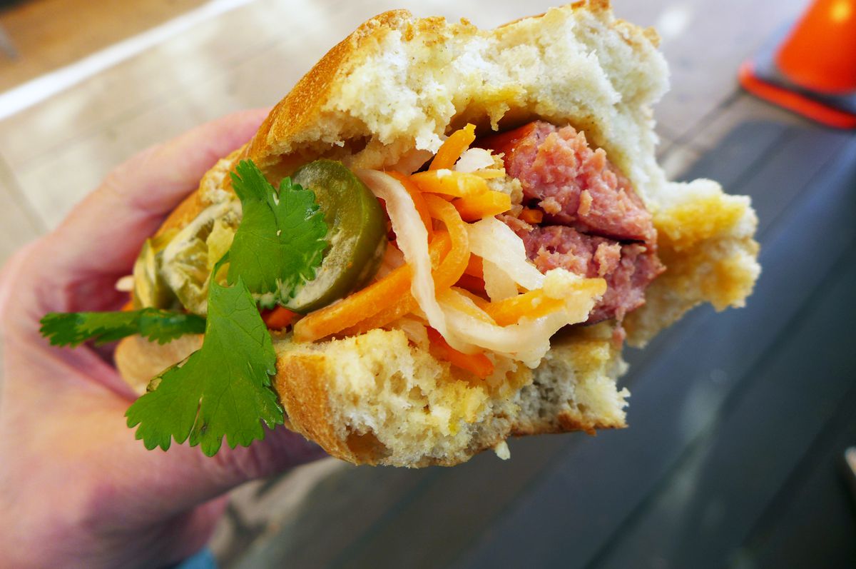 A hot dog with a bite out of one end, revealing the pink inside of a sausage, with a slaw and cilantro on top.