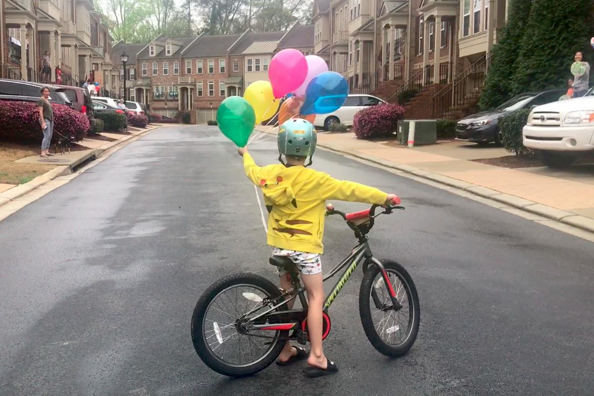 A little boy has a lot of balloons tied to his bike in a community of townhomes. 