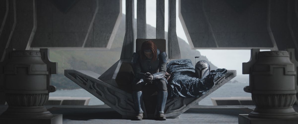 Bo-Katan (Katee Sackhoff) seated on her family’s ancestral throne, hunched over with her head held low and her helmet by her side.