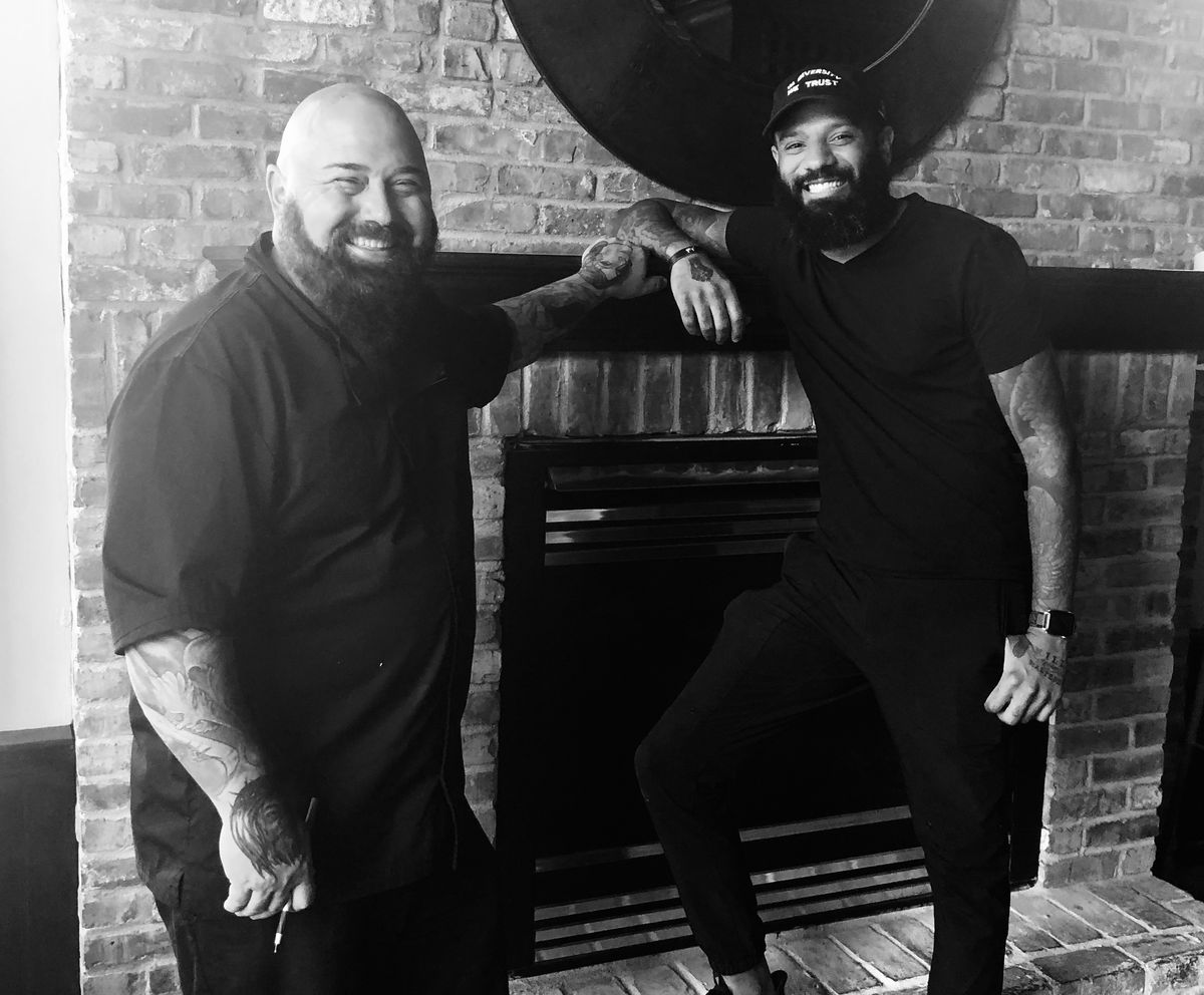 A smiling Brian Ingram and Justin Sutherland lean on a fireplace upstairs at The Gnome. Ingram is bald, bearded, and white. Sutherland is wearing his ever-present In Diversity We Trust baseball hat, clad in all black, and Black