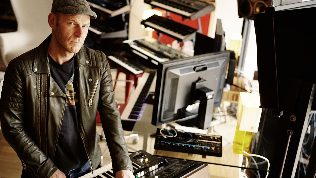 A man wearing a cap and a leather jacket is surrounded by keyboards and synthesizers.