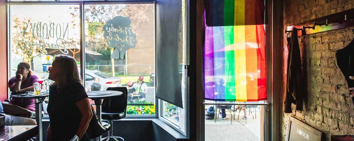 A rainbow flag hangs over the glass door of a narrow bar