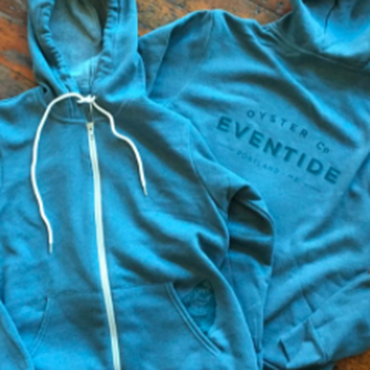 Two turquoise lightweight zip-up hoodies sit on a wooden floor
