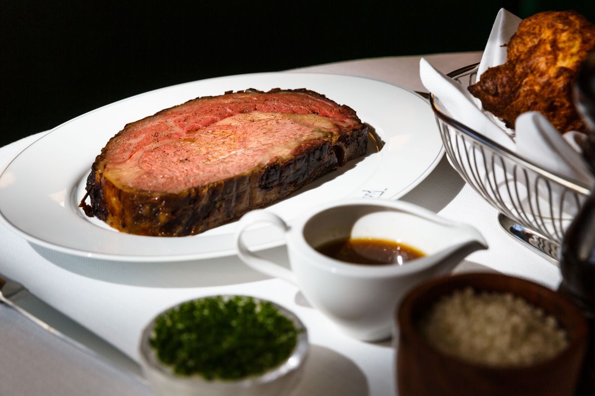 A large, pink prime rib sits on a white plate, with small side dishes in the foreground