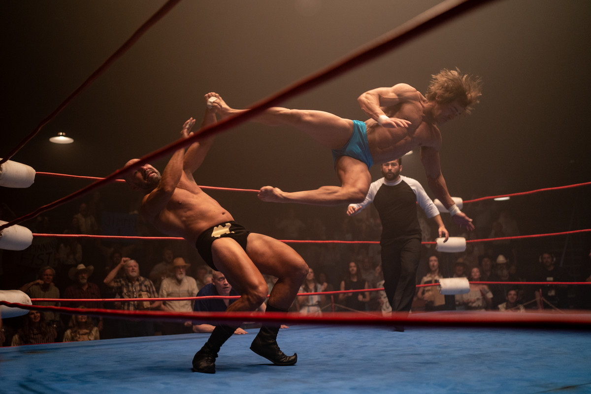 Zac Efron delivers a flying kick in a wrestling ring in The Iron Claw, very high above the ground with his hair flying.