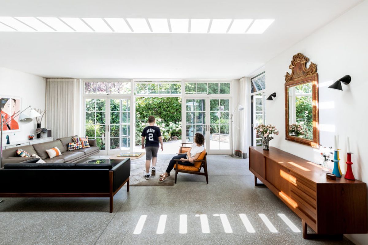 Shot of white-walled living room with large full-height steel-framed windows looking onto yard and terrazzo floors and Scandinavian furniture. 