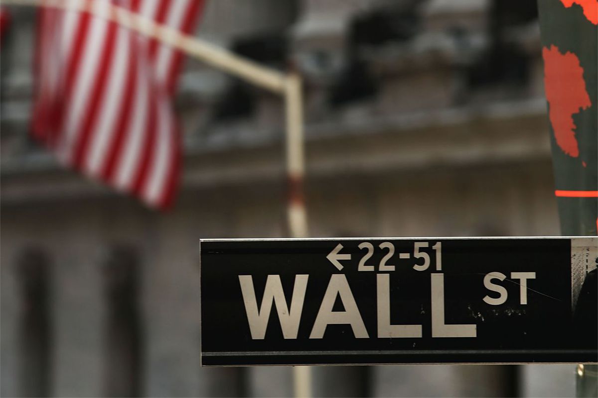 A street sign that reads “Wall St” with a building and an American flag in the background