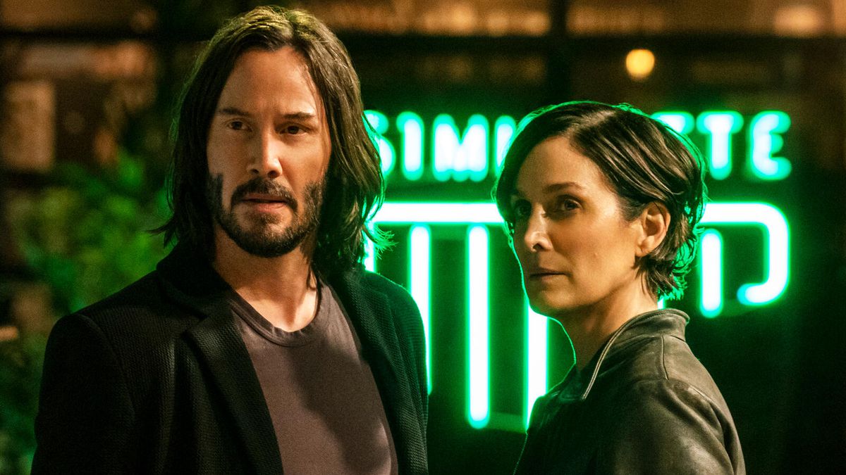 Keanu Reeves and Carrie-Anne Moss look back at the camera, a glowing neon sign behind them.