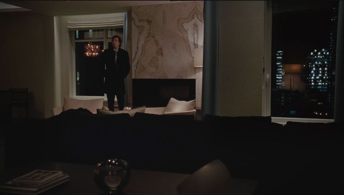 A man stands in a large living room in the HBO show Succession. The wall behind him is marble. The room is dimly lit and there are floor to ceiling windows with a view of city buildings.