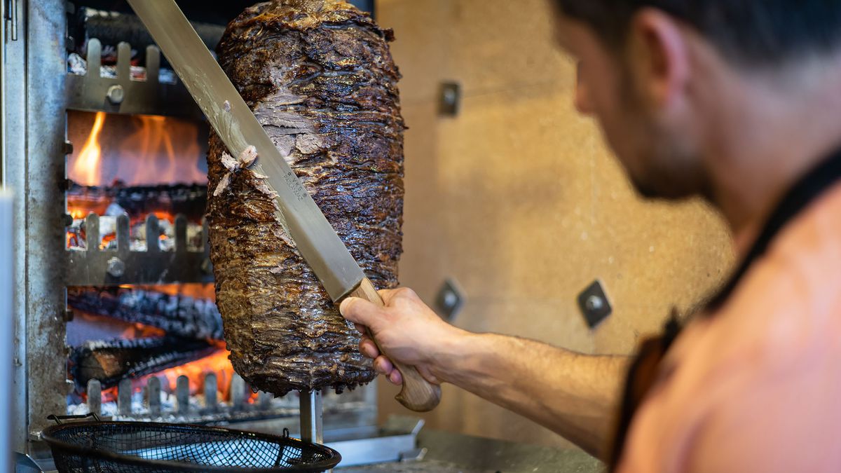 A worker with a long knife shaves meat from a vertical spit.