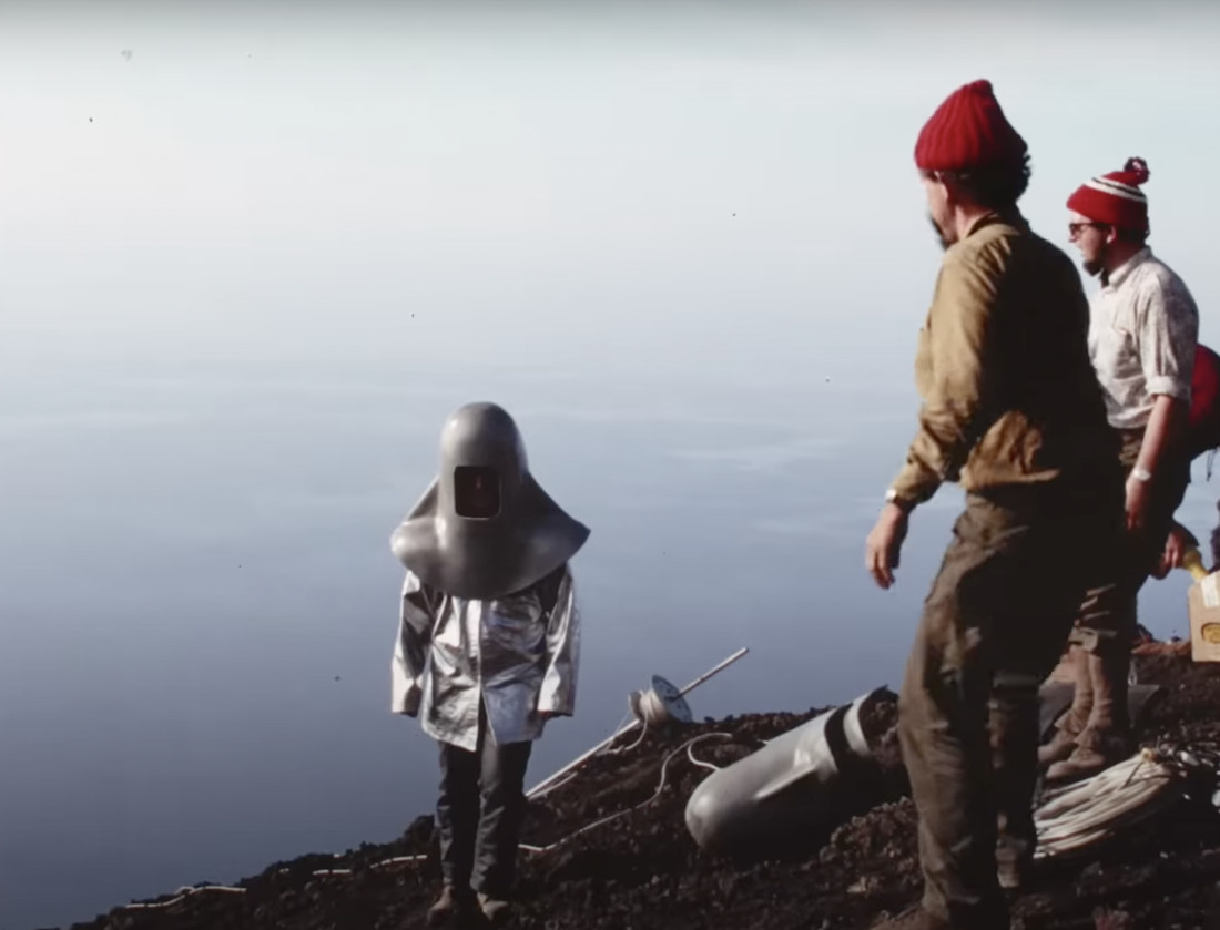 Two men look at a figure in a silver volcano suit.