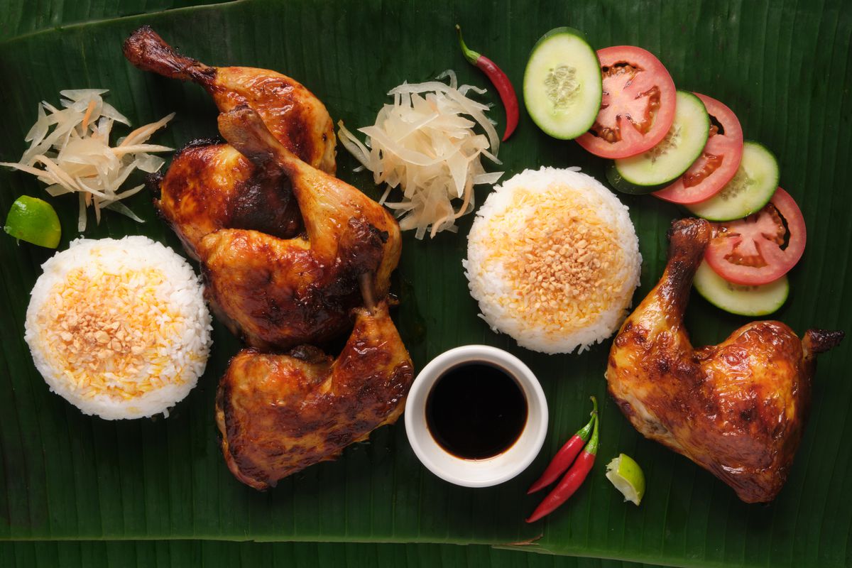 An overhead shot of chicken inasal, served with white rice, dipping sauce, and sliced tomatoes and cucumbers.