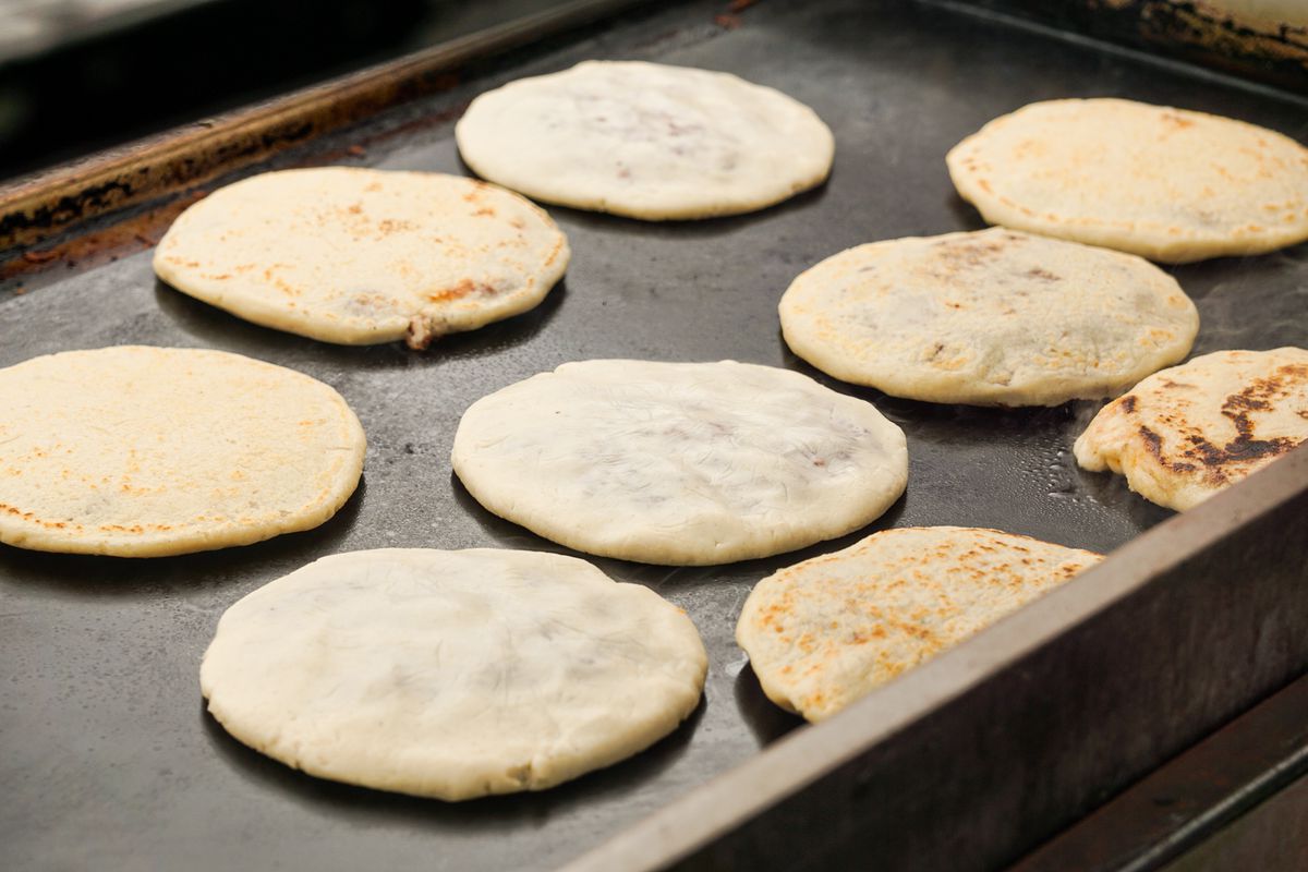 A warm griddle holds nine round masa discs to make pupusas, with some already griddled, at a daytime street vendor market in Los Angeles.