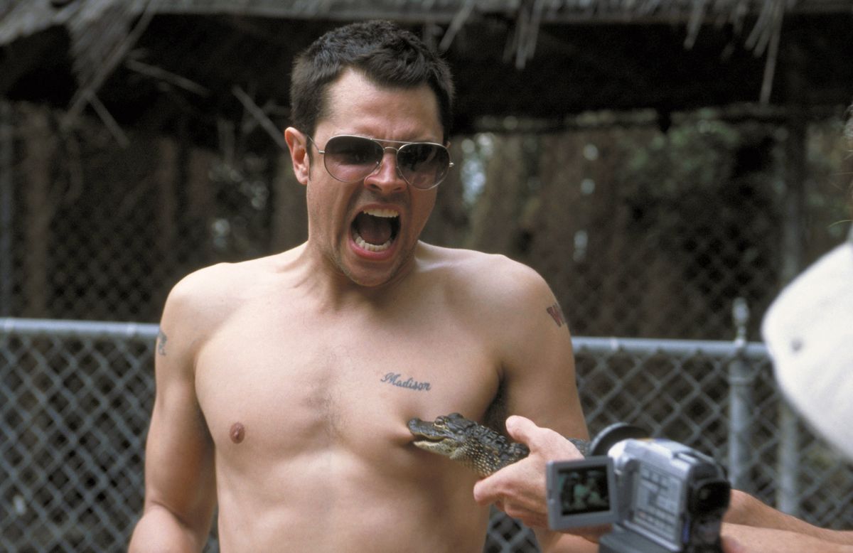 A shirtless white man in sunglasses yells as a small alligator clamps directly onto his left nipple.