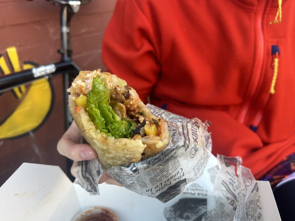 A person wearing a red sweater clutches a burrito overflowing with bulgogi.