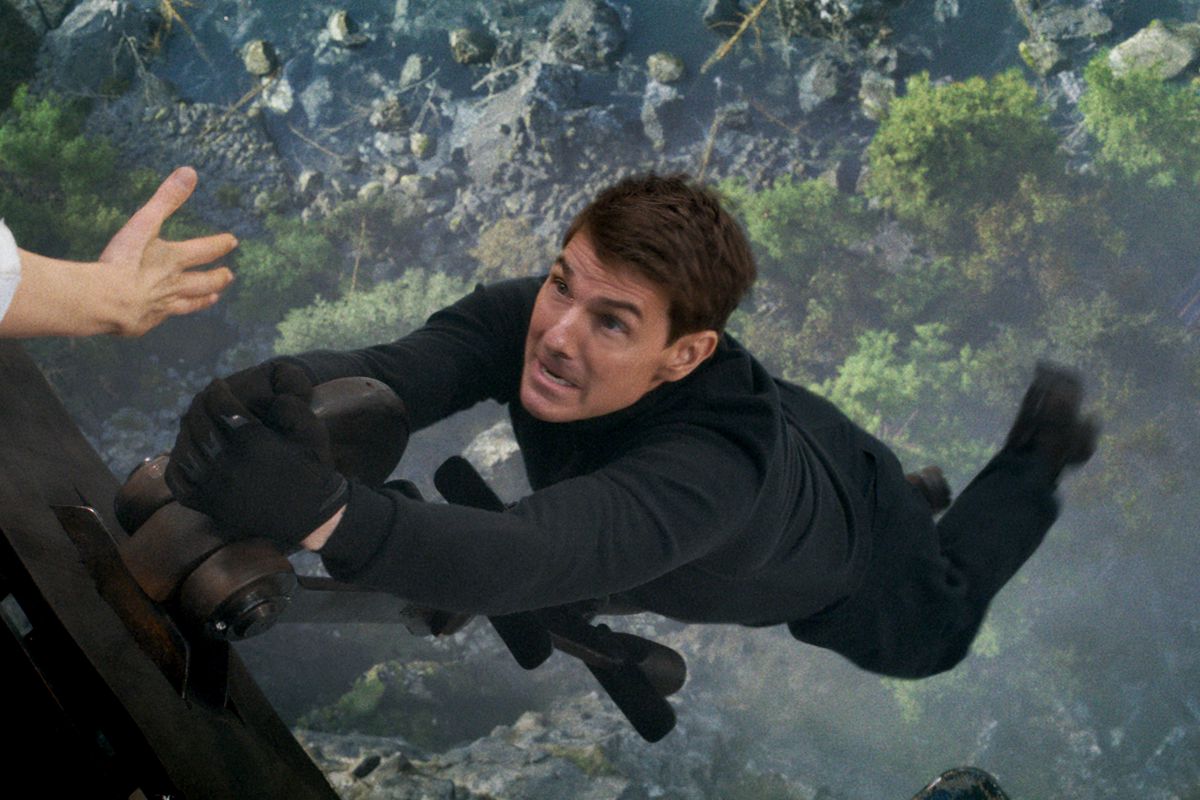 Tom Cruise as Ethan Hunt hanging off the edge of a train with a ravine very far below him as an outstretched hand from someone offscreen reaches for him in Mission: Impossible — Dead Reckoning Part 1