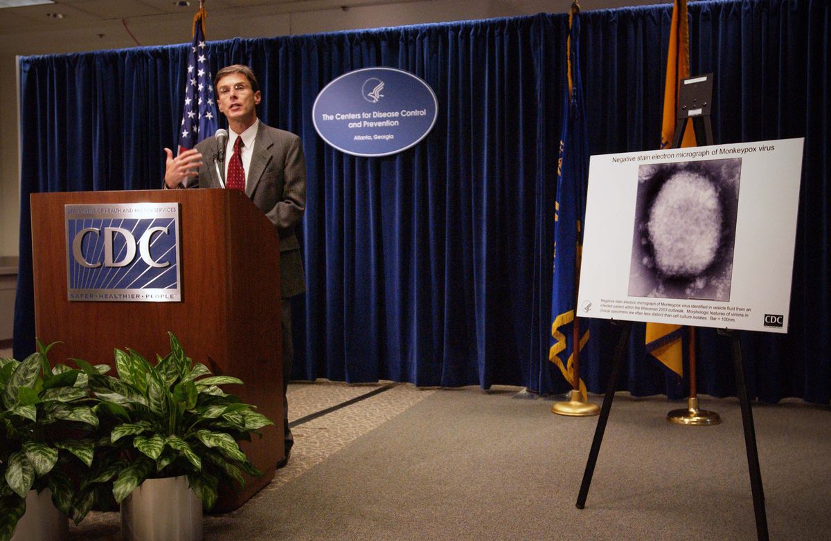 A man stands in front of a podium with the CDC logo on it.  Behind him hangs a poster on a blackboard showing a monkeypox virus under a microscope.