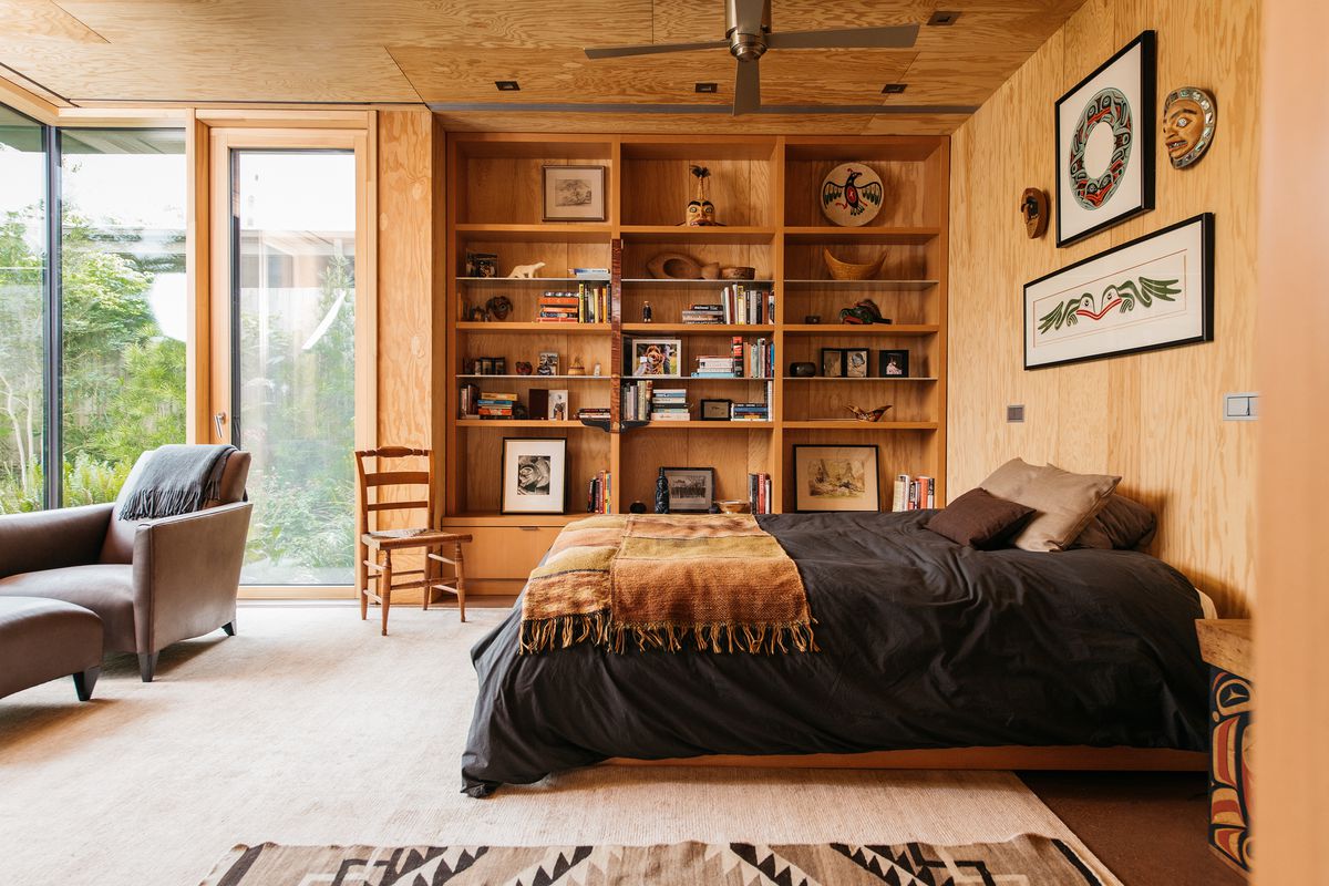 A bedroom. There is a large bed with black bed linens and a gold colored blanket. Along the far wall are shelves with various object. Art hangs above the bed. There is a tan colored area rug under the bed. Another wall has floor to ceiling windows.