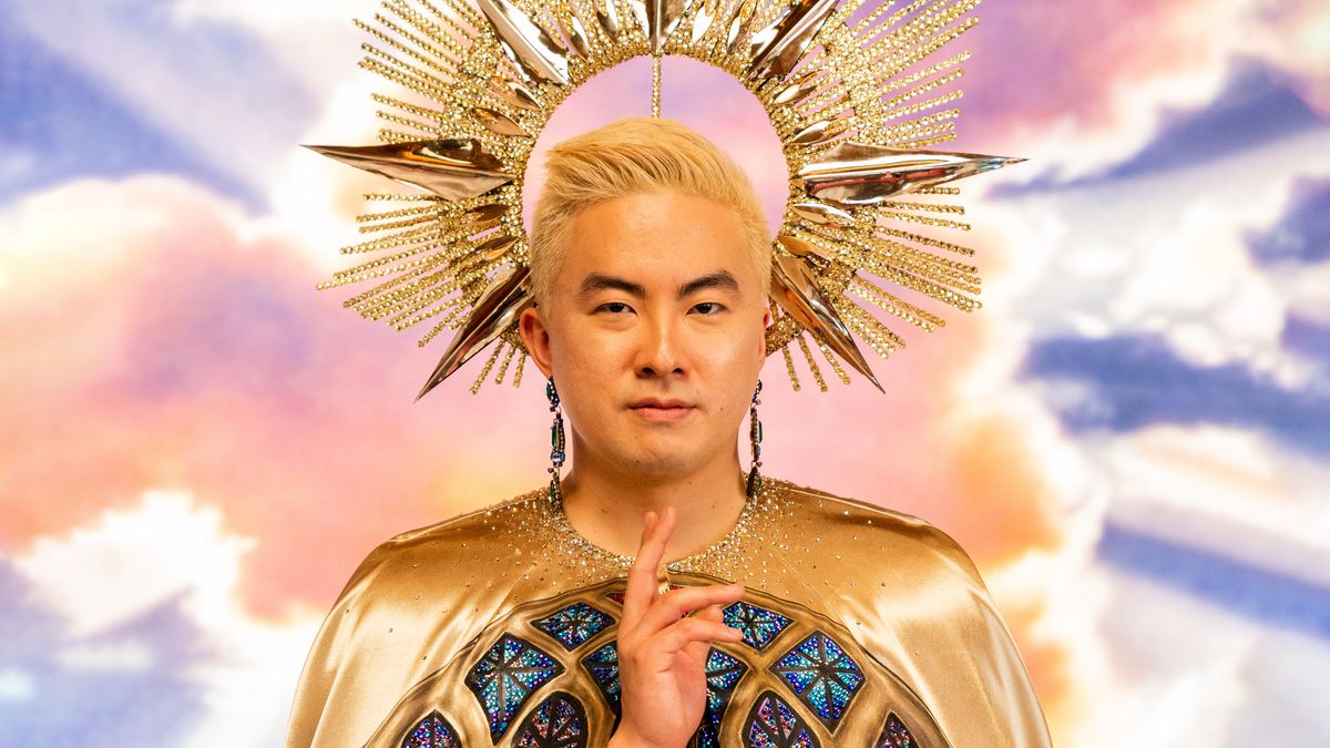 Bowen Yang as God in Dicks: The Musical, a blonde-haired man standing among clouds with a stylized sunburst crown above his head, and wearing ornate robes resembling stained glass