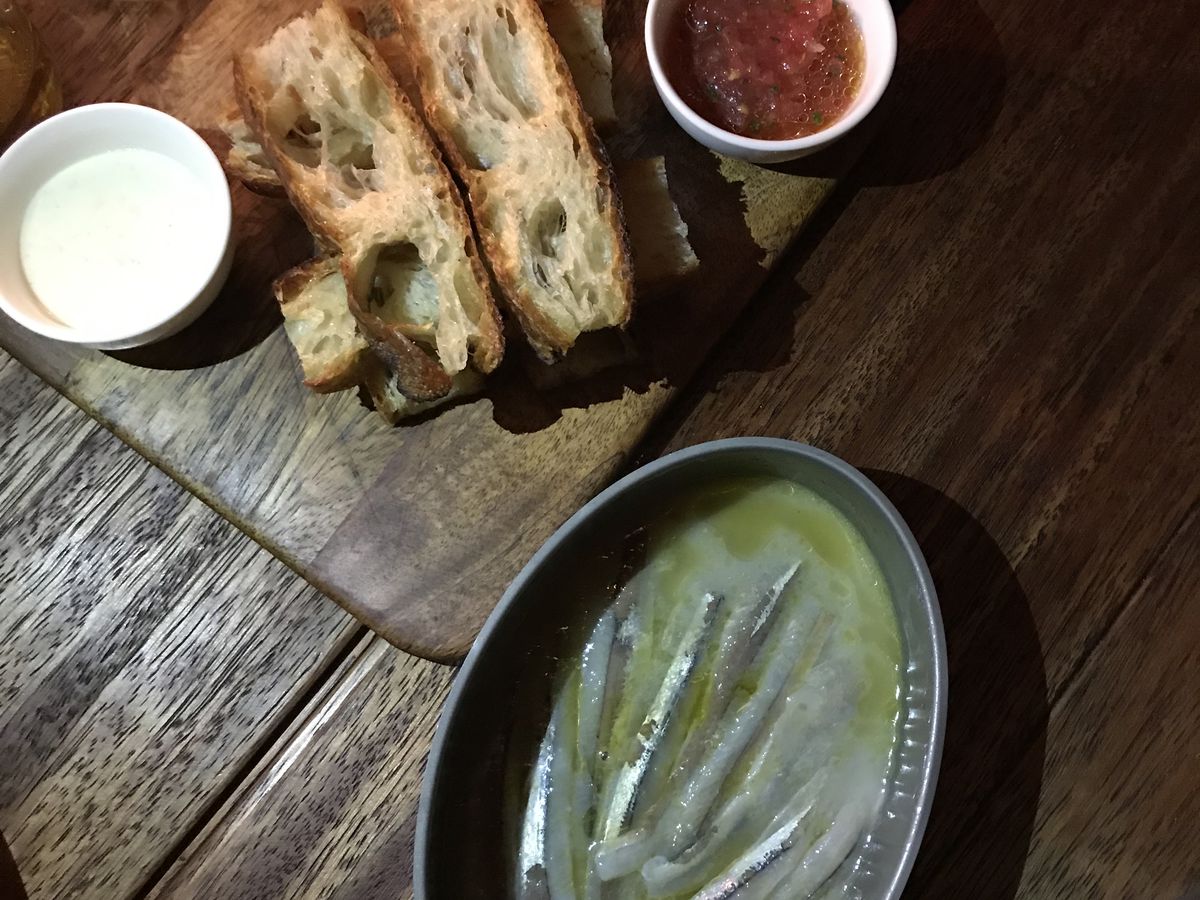 A bowl of smelt in sauce is accompanied by a bread board with slices of avocado and tomato sauce