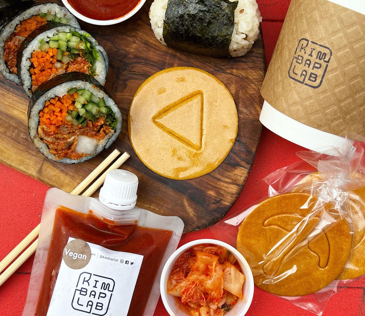 A dalgona candy with a triangle in the middle on a cutting board surrounded by kimbap, a container of kimchi, a coffee cup, chopsticks, and gochujang.