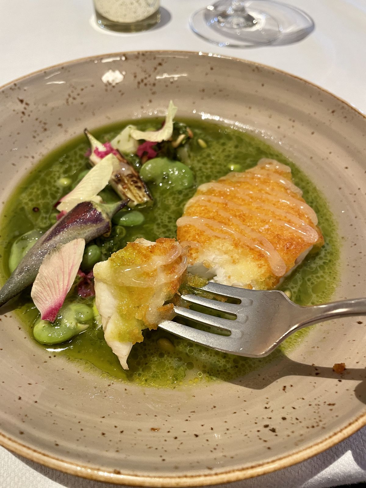 A bowl holds a filet of snapper that has been crusted and sits in an arugula sauce. There are also oversized lima beans and radishes in the dish. A fork holds a bite.