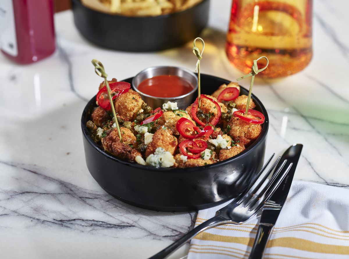A black bowl with crispy-fried cauliflower topped with sliced red chiles, blue cheese crumbles, and a side of hot sauce on a marble table with drinks, napkin, and cutlery.