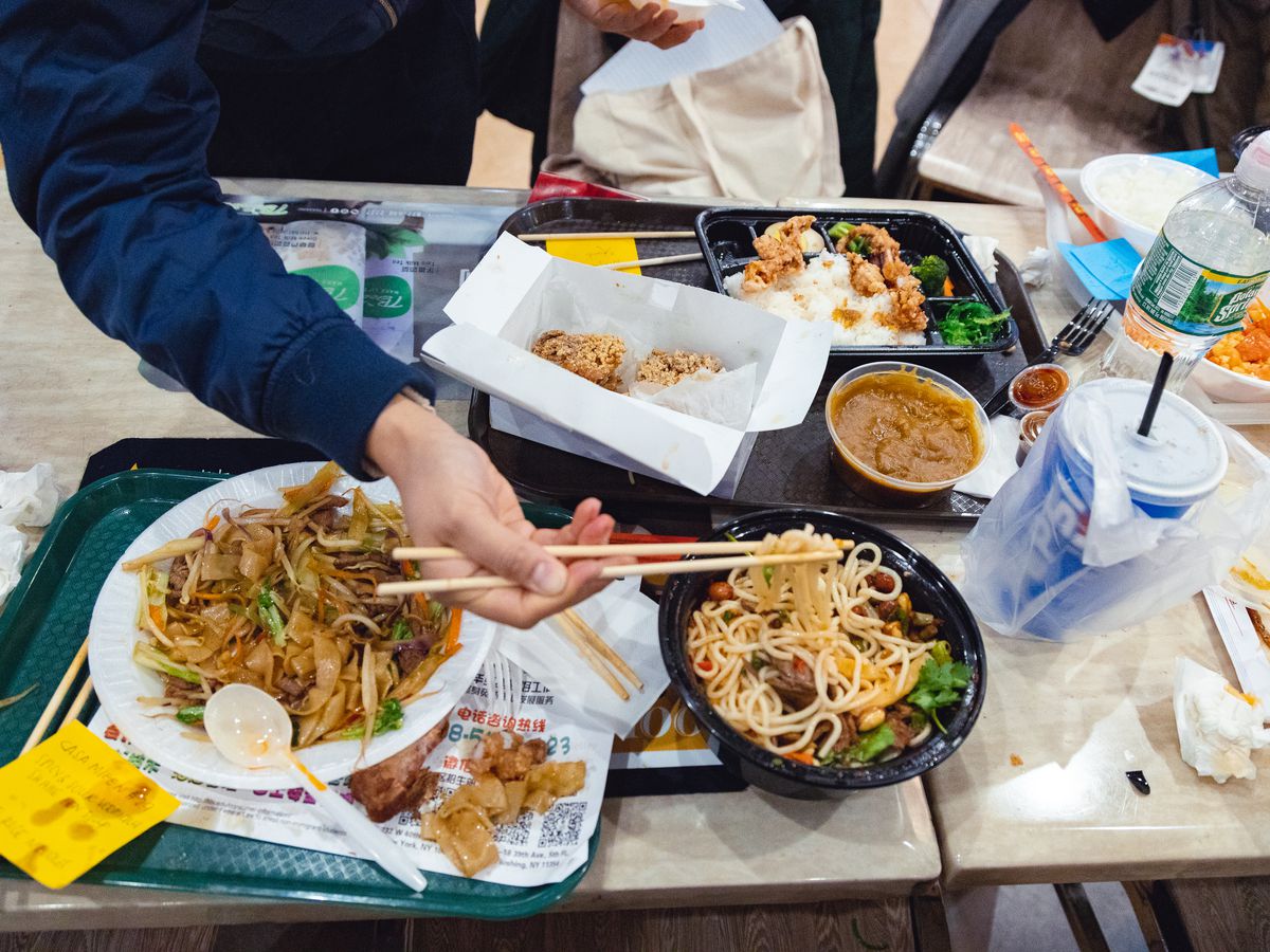 A hand holds a noodle with chopsticks over a to-go bowl.