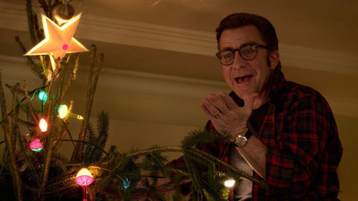 A man wearing glasses (Peter Billingsley) stands on a ladder next to a Christmas tree, looking exasperated with his hand raised and mouth open.