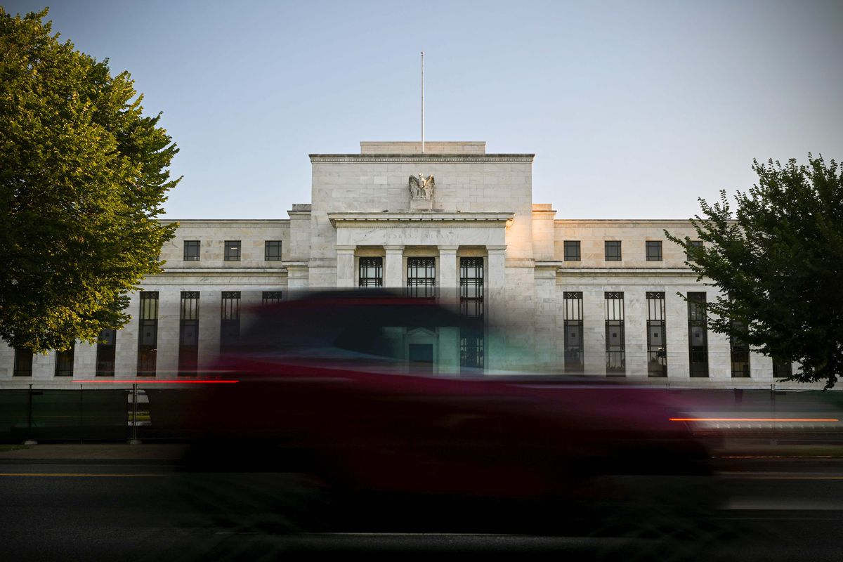 A time-lapse photo shows the Federal Reserve building, a white stone-fronted building with tall columns and vertical windows, as a blurred stream of traffic passes in front of it and the sunlight on the surrounding trees moves slowly.