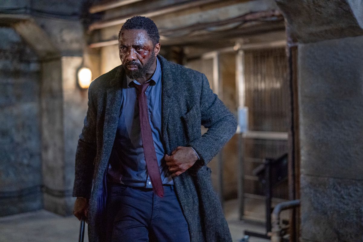 A man (Idris Elba) dressed in a blue dress shirt, burgundy tie, and a tweed long coat with visible bruises and blood on his face.