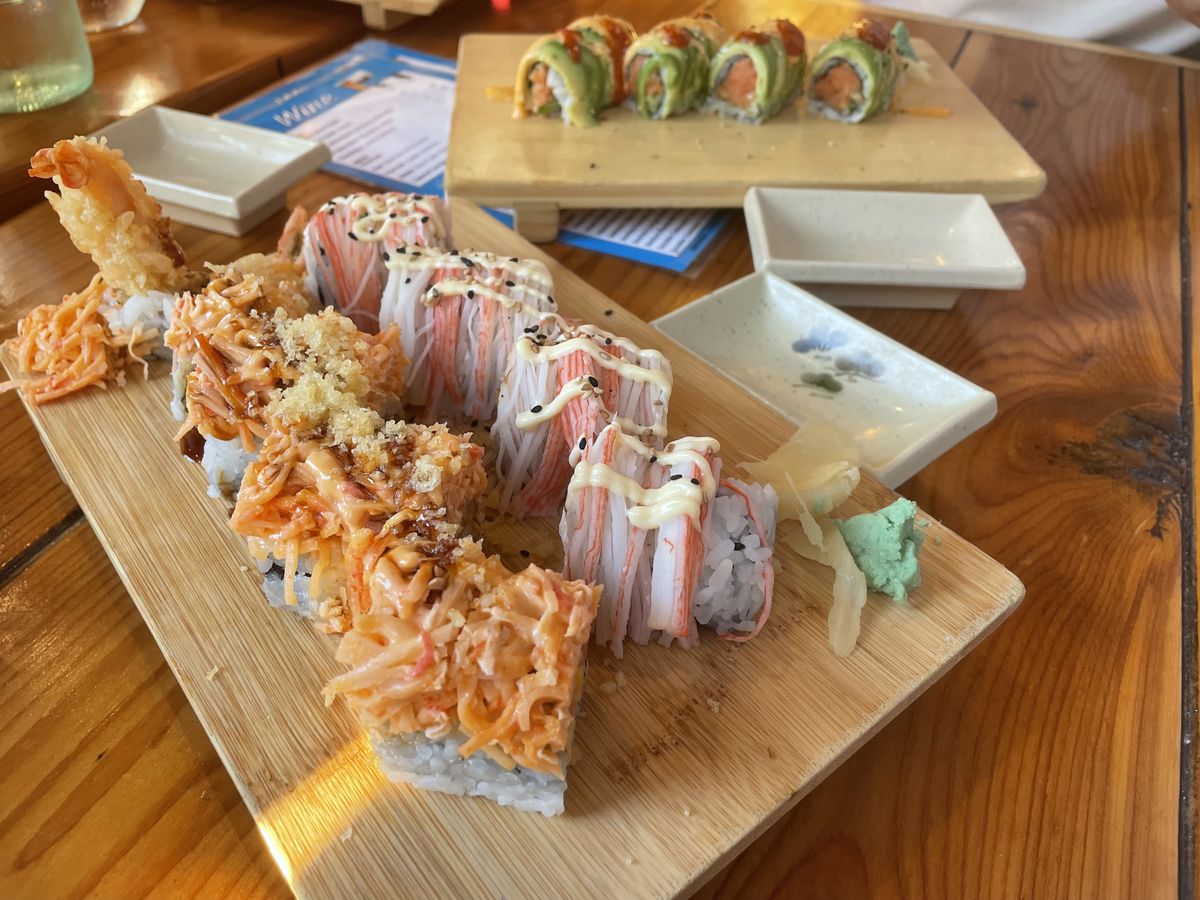 Two rolls of sushi sit on a wooden plate with another visible in the background. 