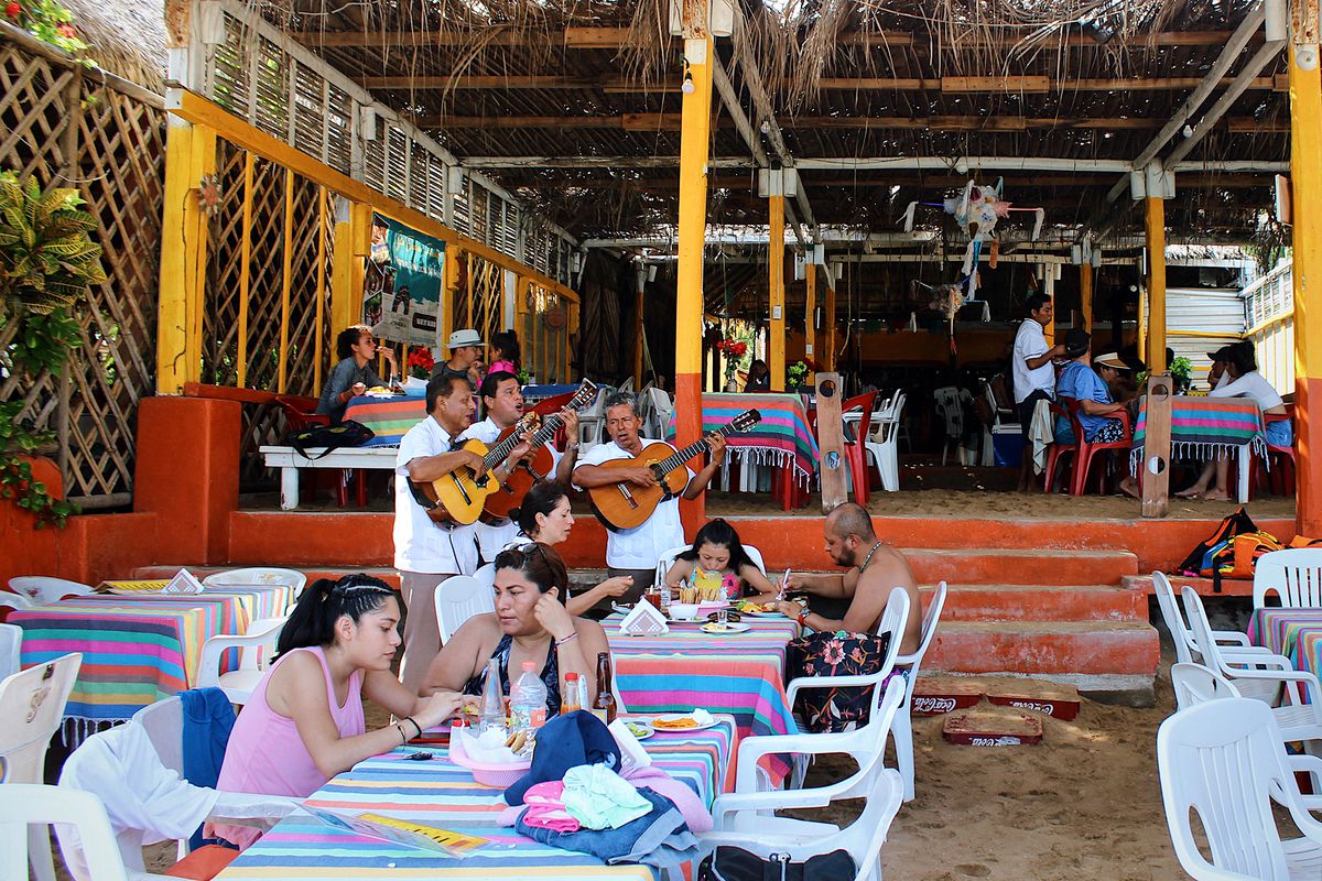 Diners eat at tables in the sand while three guitarists play in the background.