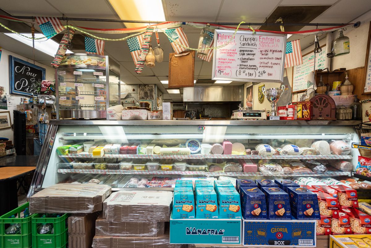 A deli counter with imported goods on the floor, meat at the ready, and hanging signs.