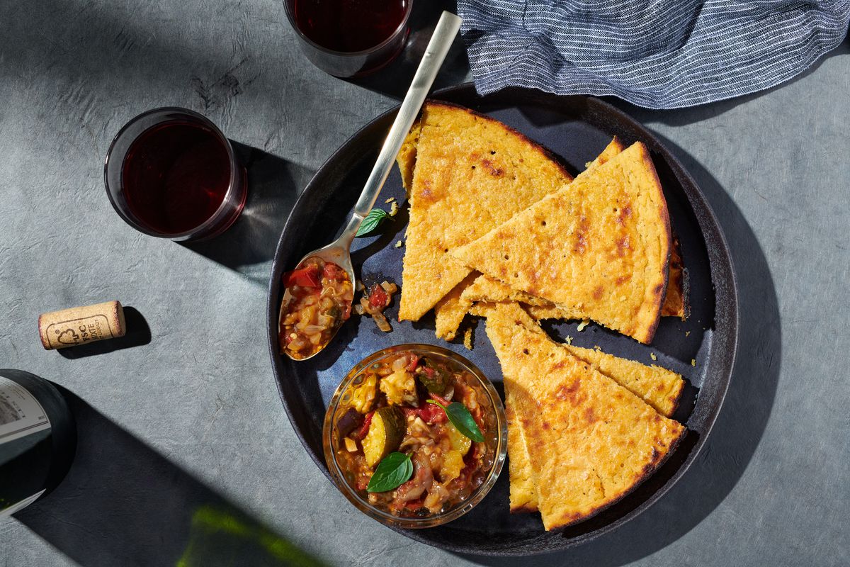 Wedges of socca, a bowl of ratatouille, and a big spoon sit on a big black plate. Next to the plate, on a grey tabletop, there are two glasses of red wine, a striped blue napkin, a cork, and a wine bottle.