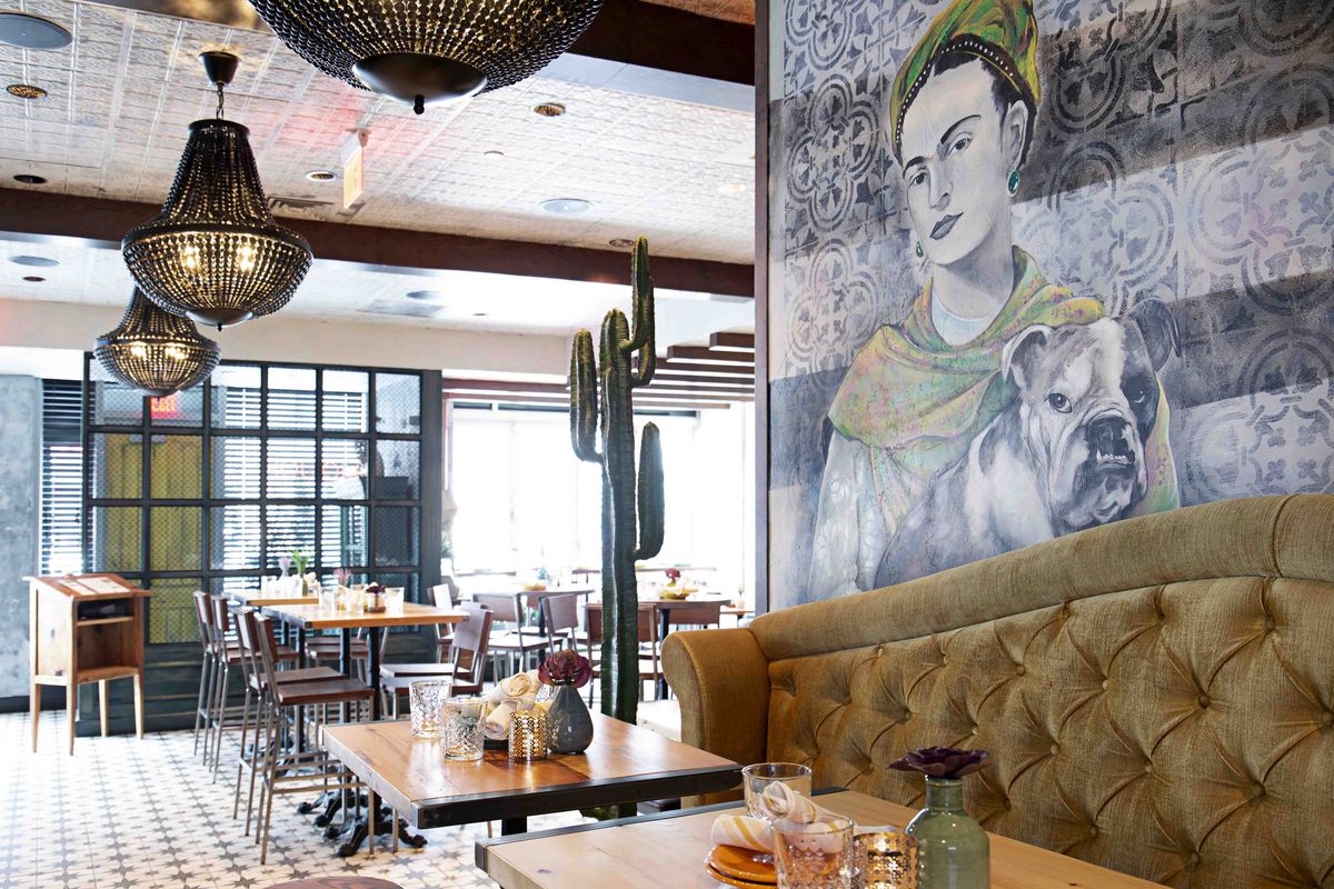 Wide angle of a restaurant with a tufted, velvet banquette in the foreground, wooden tables and chairs in the rear, and a wall mural featuring Frida Kahlo with a bulldog behind the banquette