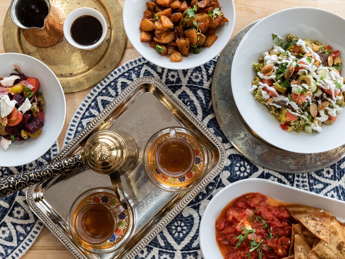 A Moroccan tea set, a red dip with pita chips, a lamb tagine, harissa potatoes, coffee, and more on a wooden table with blue and white-patterned woven center pieces.