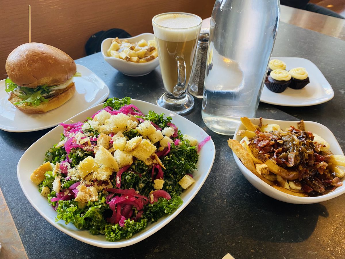 A big bowl of kale salad, a breakfast sandwich, two bowls of poutine, a latte, and some cupcakes