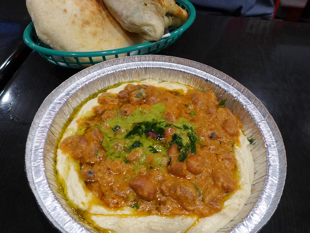 A round container of fluffy hummus with deep brown fava beans and a green sauce on top.