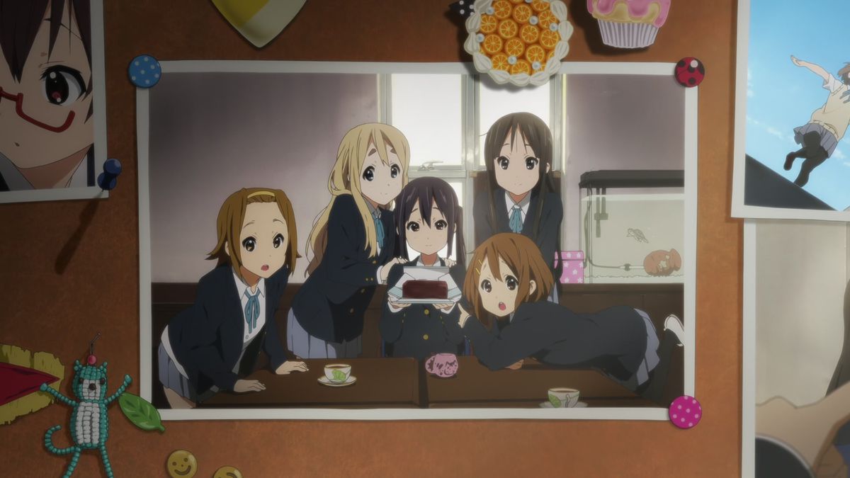 A group of five school girls — the main characters of K-ON! — pose around a table. The girl in the middle, who has black pigtails, is holding a cake. The photo of the girls is pinned onto a bulletin board.