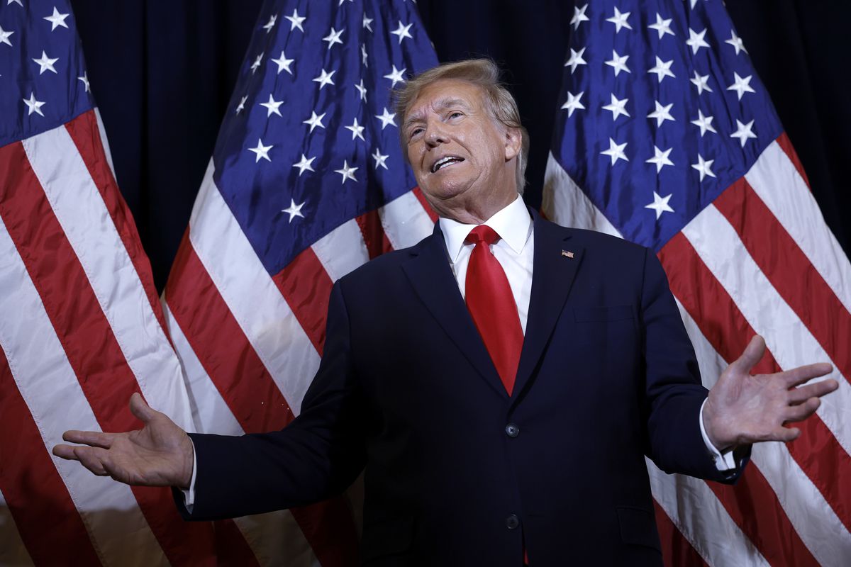 Donald Trump stands before three American flags with arms outspread, mid-speech.