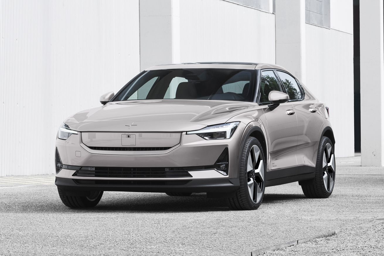 a silver and beige colored polestar 2 is parked on concrete with a white building in the back. The front and drivers side of the car are visible with a new front that has a body colored panel covering the grille.