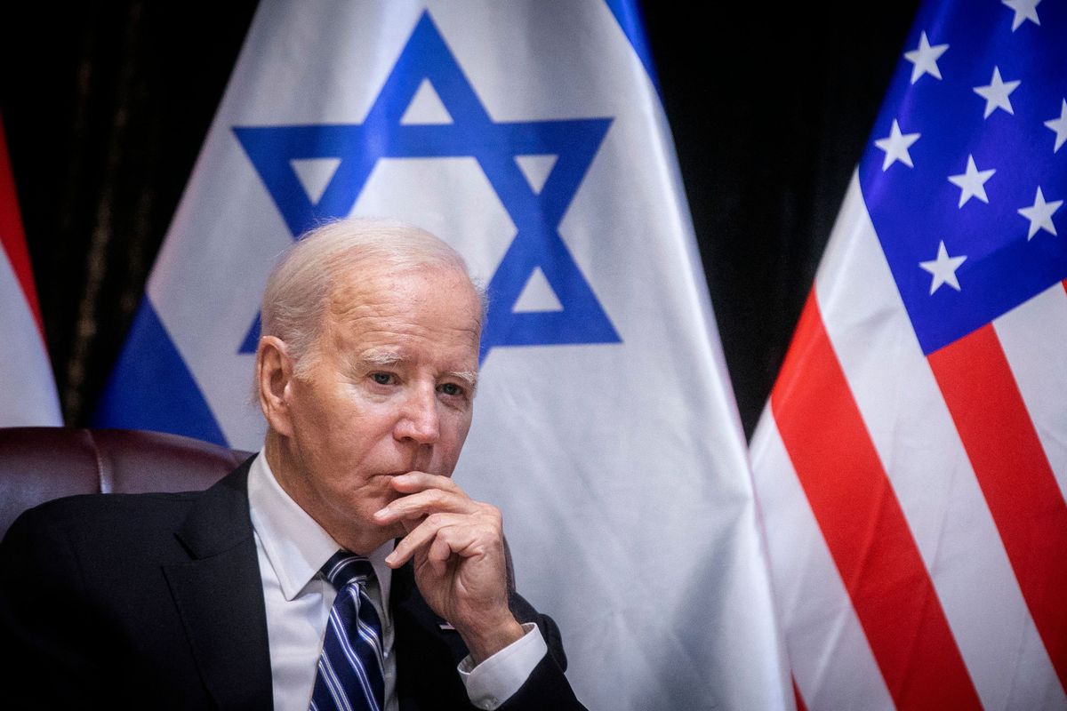 Biden sits in a chair with the Israeli and US flags in the background.
