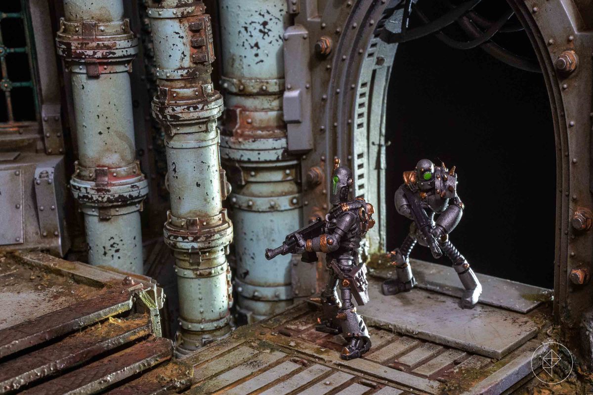 Two automatons step out of the darkness and into the bridge of a gothic spaceship. Their solitary eyes glow, primitive rifles held in their hands. The background is rotting, rusted, and flowing ominously read.