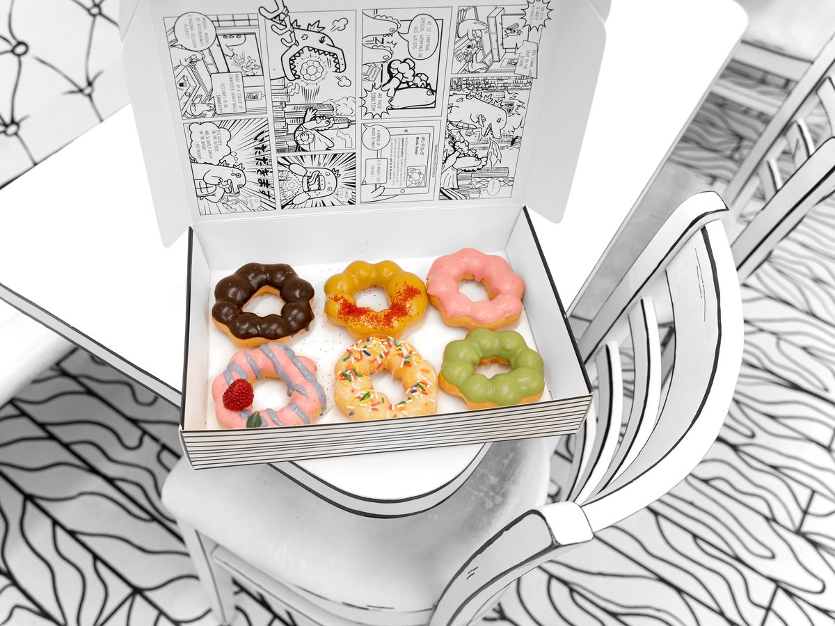 Six colorful mochi doughnuts sit inside an black and white illustrated box in a black and white cafe space.