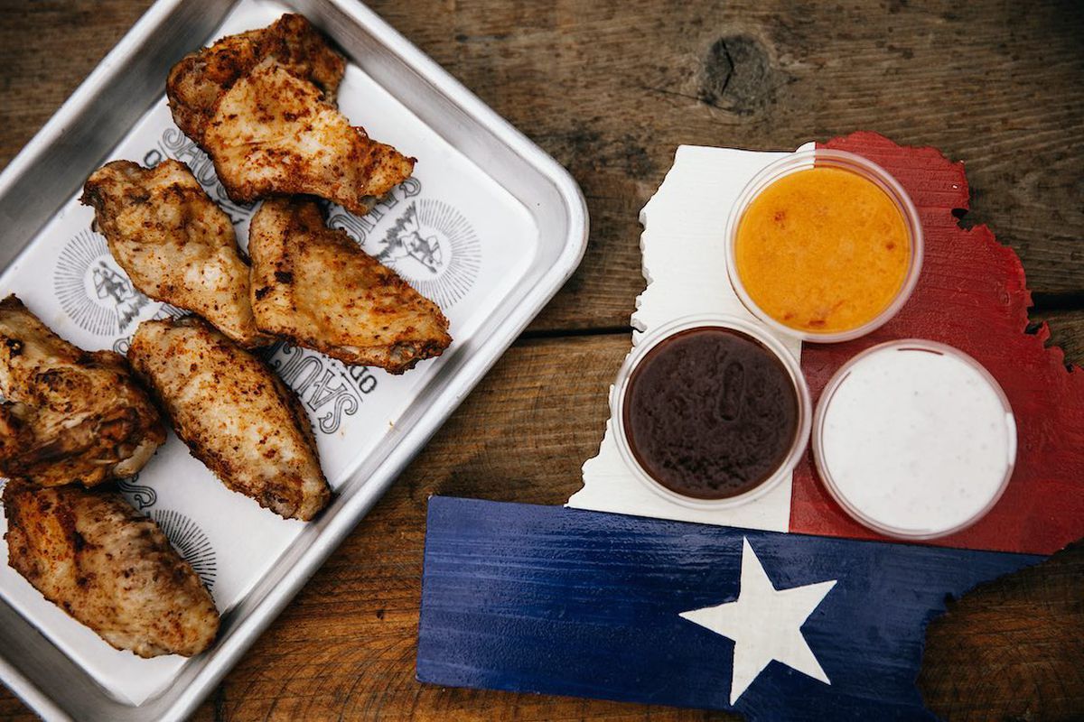 A tray of chicken wings next to a wooden tray painted to look like the Texas flag with three sauces in round containers