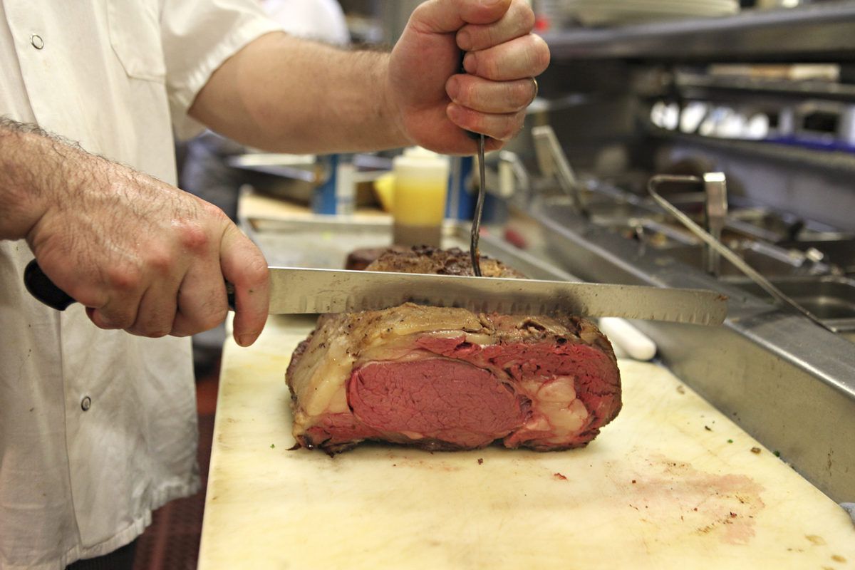 A chef carves a giant hunk of prime rib on a white butchers block. 