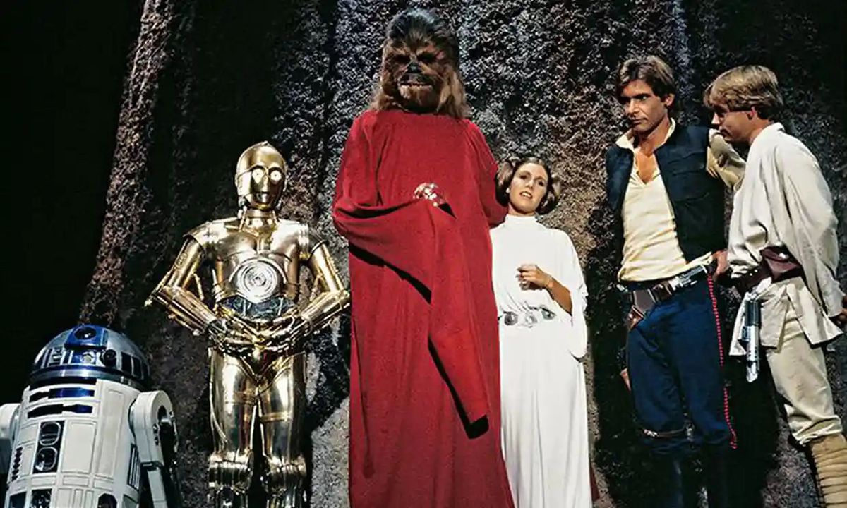 A stock publicity photo from the 1978 Star Wars Holiday Special shows R2-D2, C-3PO, Chewbacca (in a long red robe), Carrie Fisher as Princess Leia, Harrison Ford as Han Solo, and Mark Hamill as Luke Skywalker, all standing together in front of a tall, grainy stone